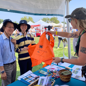 Tabling2 Young woman in a ball cap hands an orange Park People bag to two festival goers at a Park People table