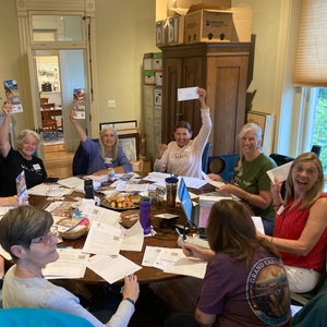 Six women sit at a round table, writing letters and showing their letters to the camera with big smiles