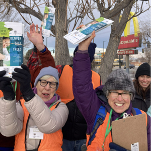 Group of women in hats and coats in front of a McDonalds sign, holding up Denver Digs Trees doorhangers. They're wearing safety vests and have big smiles.