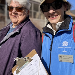 Two women standing in the sunshine, wearing sunglasses. One wears a blue Community Forester vests, and is holding a clipboard with a site inspection form